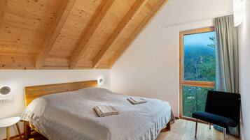 Ein Schlafzimmer mit Holzbalkendecke, einem großen Fenster mit Blick und einem Bett mit Decke sowie einem Sessel.