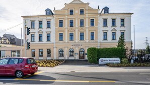 Ein großes, mehrgeschossiges Gebäude im klassischen Stil, mit einem markanten Eingang mit Stufen, einem Schild, das es als Hotel kennzeichnet, und einer kleinen Grünfläche davor.