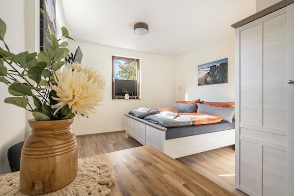 A bedroom with a bed, a wooden table with a vase, a window with blinds, and a door.