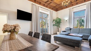 Ein modernes Wohnzimmer mit Holzboden, einem Flachbildfernseher, einem grauen Sofa, einem Esstisch mit Stühlen und großen Fenstern.