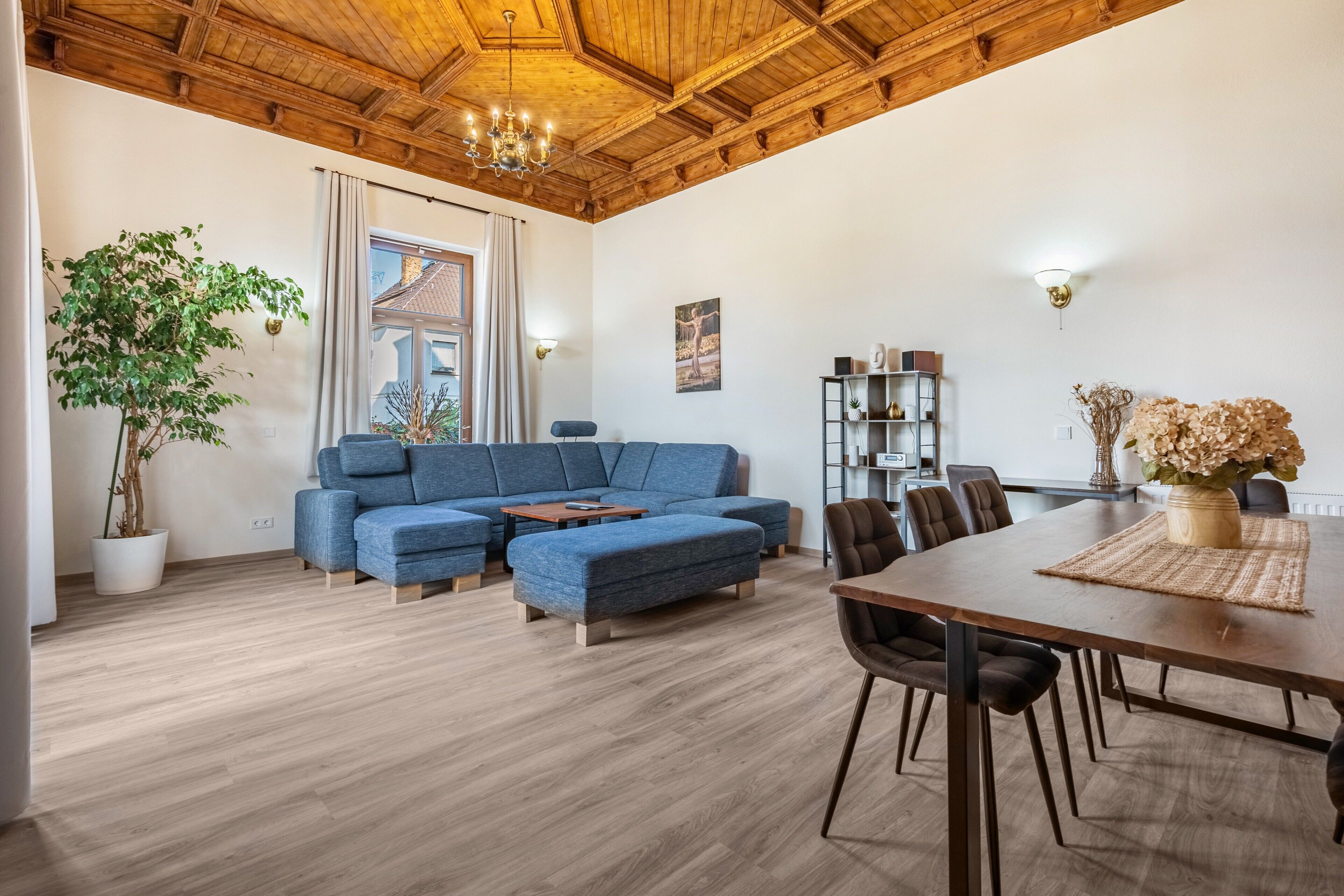 En moderne stue med en blå hjørnesofa, et spisebord med stole, en lysekrone, en stor stueplante og udsigt udad.