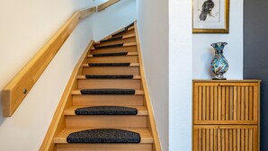 Eine Holztreppe mit schwarzen Teppichstufen, ein gerahmtes Vogelbild und eine dekorative Vase auf einem Schrank.