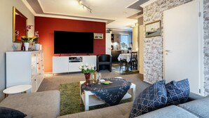 Ein Wohnzimmer mit Flachbildfernseher, einem weißen Schrank, einem Esstisch und einer Küche im Hintergrund.