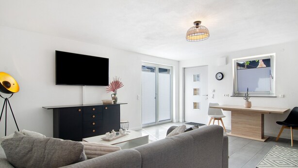 Ein modernes Wohnzimmer mit einer Couch, einem Fernseher, einer schwarzen Kommode und einem Schreibtisch mit Stuhl.