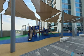 Een speelterrein met speeltoestellen en schaduwrijke zones, voor een modern gebouw.