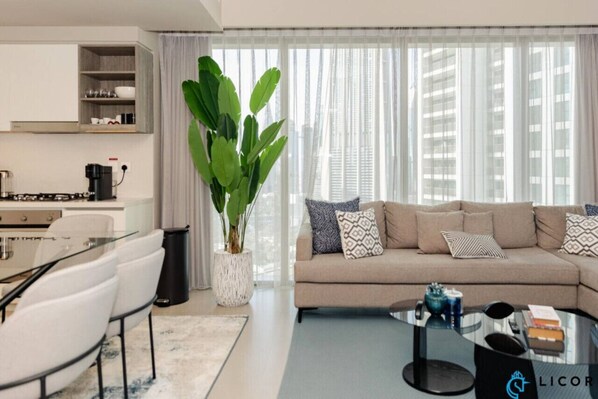 Una sala de estar moderna con sofá, mesa de centro y sillas. Hay una planta grande en una maceta blanca, y un área de cocina con armarios y electrodomésticos.
