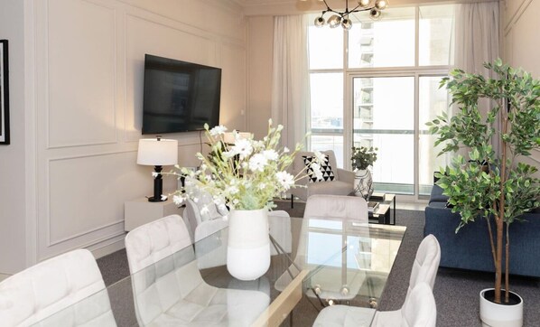 A modern living room with a glass coffee table, white chairs, a sofa, a TV mounted on the wall, and a large window with curtains. - Prime Spacious 2 BR Apt, Burj Khalifa & Canal View (Dubai)