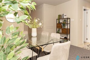 A dining area with a glass table surrounded by chairs, a white vase with flowers, and a bookshelf with various items. - Prime Spacious 2 BR Apt, Burj Khalifa & Canal View (Dubai)