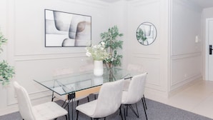 A modern dining area with a glass table, white chairs, a vase with flowers, and a framed abstract painting. - Prime Spacious 2 BR Apt, Burj Khalifa & Canal View (Dubai)