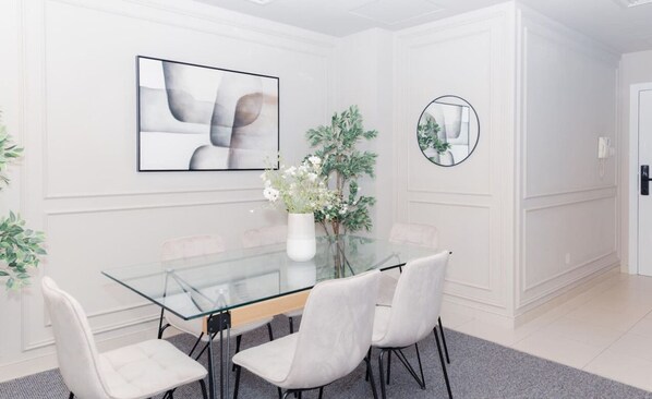 Un espace de salle à manger moderne avec une table en verre, des chaises blanches, un vase rempli de fleurs et un tableau abstrait encadré.