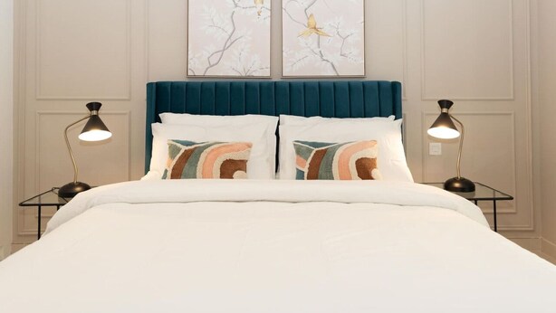 A bed with a teal upholstered headboard, two bedside lamps, and two wall-mounted paintings. - Prime Spacious 2 BR Apt, Burj Khalifa & Canal View (Dubai)