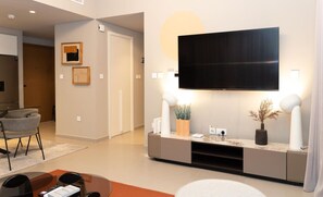 Una sala de estar moderna con un televisor de pantalla plana montado en la pared, una zona de estar con sillas, una mesa de centro y un aparador con objetos decorativos.