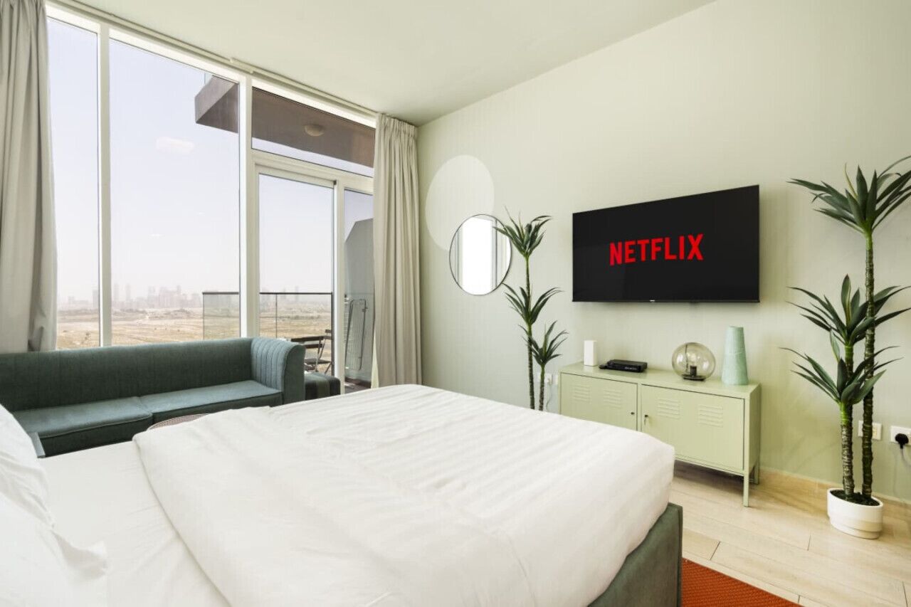 A hotel room with a large bed, a TV mounted on the wall, a sofa, a small table, and a view of the city through large windows.