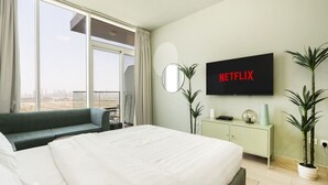 A hotel room with a large bed, a TV mounted on the wall, a sofa, a small table, and a view of the city through large windows. - Stylish Cozy Studio in Bloom Towers, Jumeirah Village Circle – Modern Comfort & City Views (Cosy Studio Apartment in Bloom Towers, Jumeirah Village Circle)