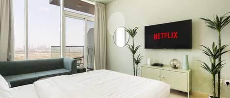 A hotel room with a large bed, a TV mounted on the wall, a sofa, a small table, and a view of the city through large windows.