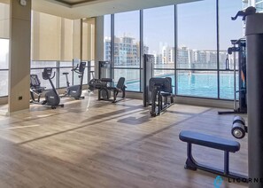 Ein Fitnessstudio mit Laufbändern, Kraftgeräten und einer Bank. Große Fenster bieten Blick auf die Stadt und ein Gewässer.