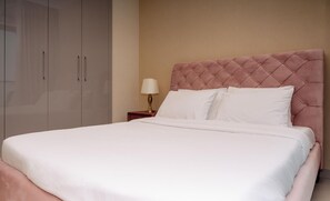 Cama bem arrumada com roupa de cama branca e cabeceira rosa, luminária de cabeceira e guarda-roupa com puxadores.