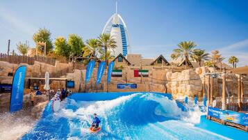 A water park with a large wave pool, a prominent building in the background, and flags.