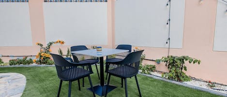 Um pátio com uma mesa redonda e quatro cadeiras, rodeado por um gramado bem cuidado e uma cerca decorativa.