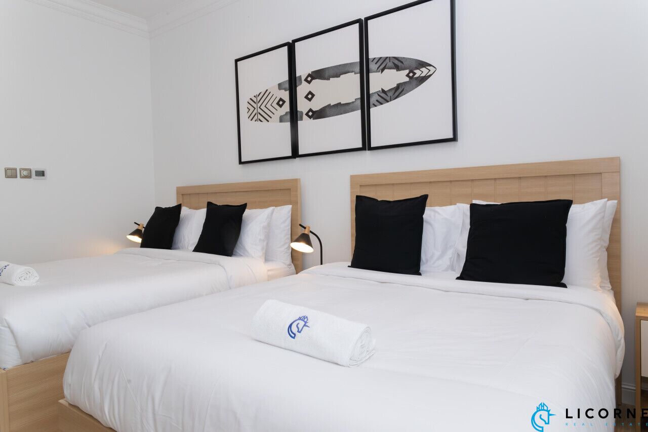 A hotel room with two beds, each with white bedding and black pillows. There is a framed artwork above the beds and a small table with a lamp beside the beds.