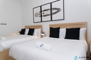 A hotel room with two beds, each with white bedding and black pillows. There is a framed artwork above the beds and a small table with a lamp beside the beds.
