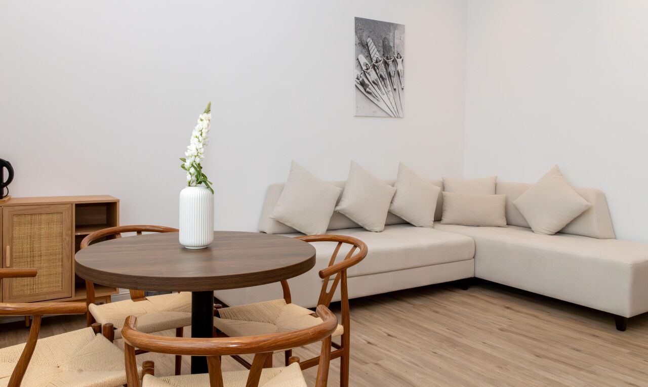 A modern living room with a round wooden table, beige sofa, and a white vase with flowers.