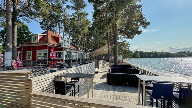 Una terraza de madera con mobiliario exterior y un edificio rojo con techo inclinado.