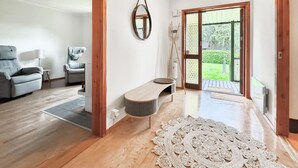 Una sala de estar moderna con un sofá gris, una silla, una mesita y un banco de madera. Hay una alfombra grande de fibras naturales en el suelo y una puerta de madera que da a un jardín.