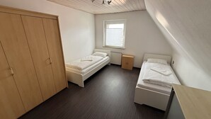 Pieni, siisti huone, jossa on kaksi erillistä sänkyä, puinen vaatekaappi, patteri ja ikkuna, jossa on sälekaihtimet.