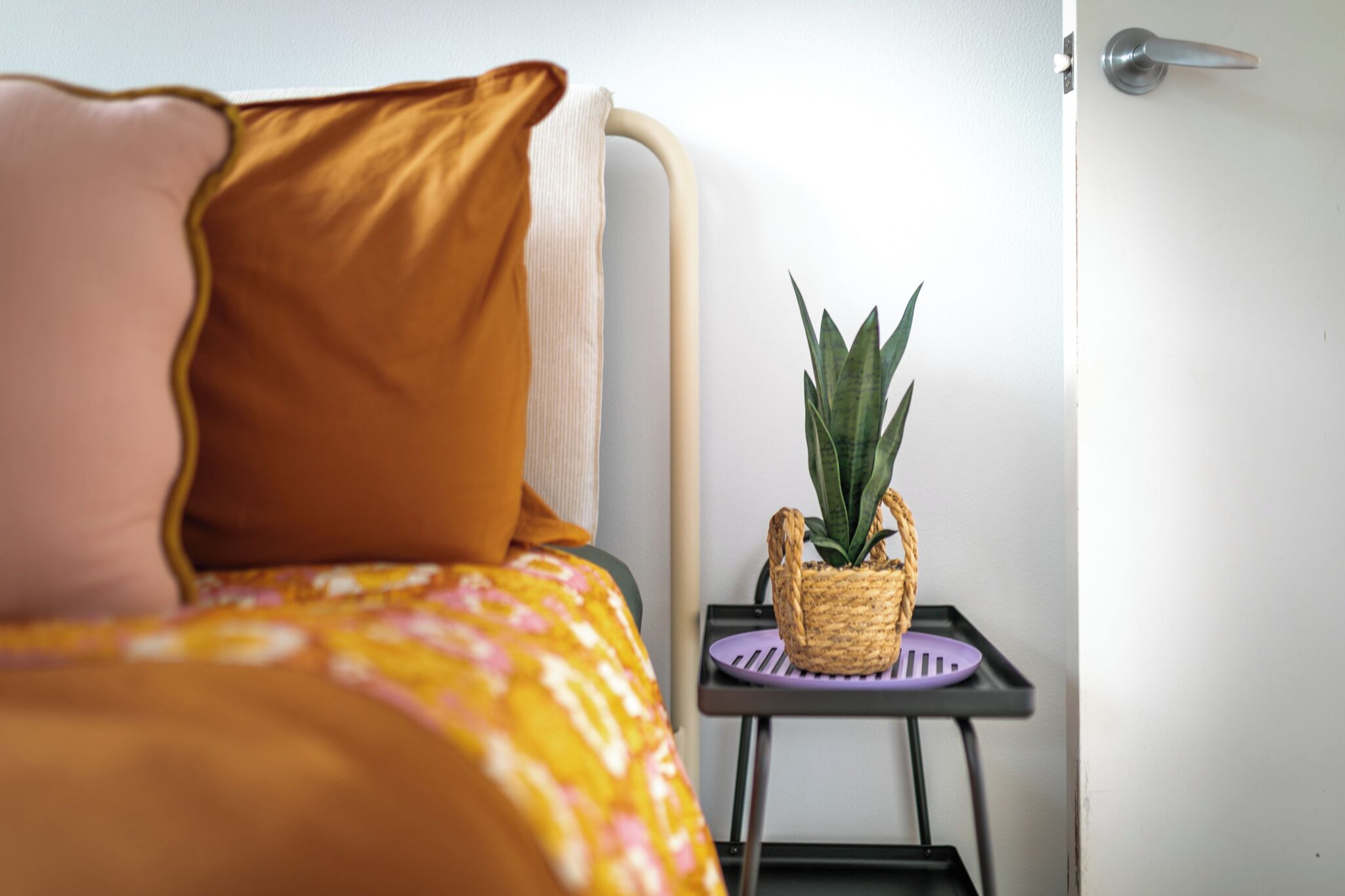 Un lit recouvert d’une couvre-lit à motifs jaunes et roses, une petite table sur laquelle est posée une plante en pot, et une porte dotée d’une poignée argentée.