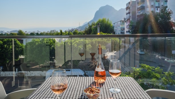 A table with a bottle and glasses on a balcony overlooking a cityscape with mountains. - Rudblu Suites (Konyaalti)
