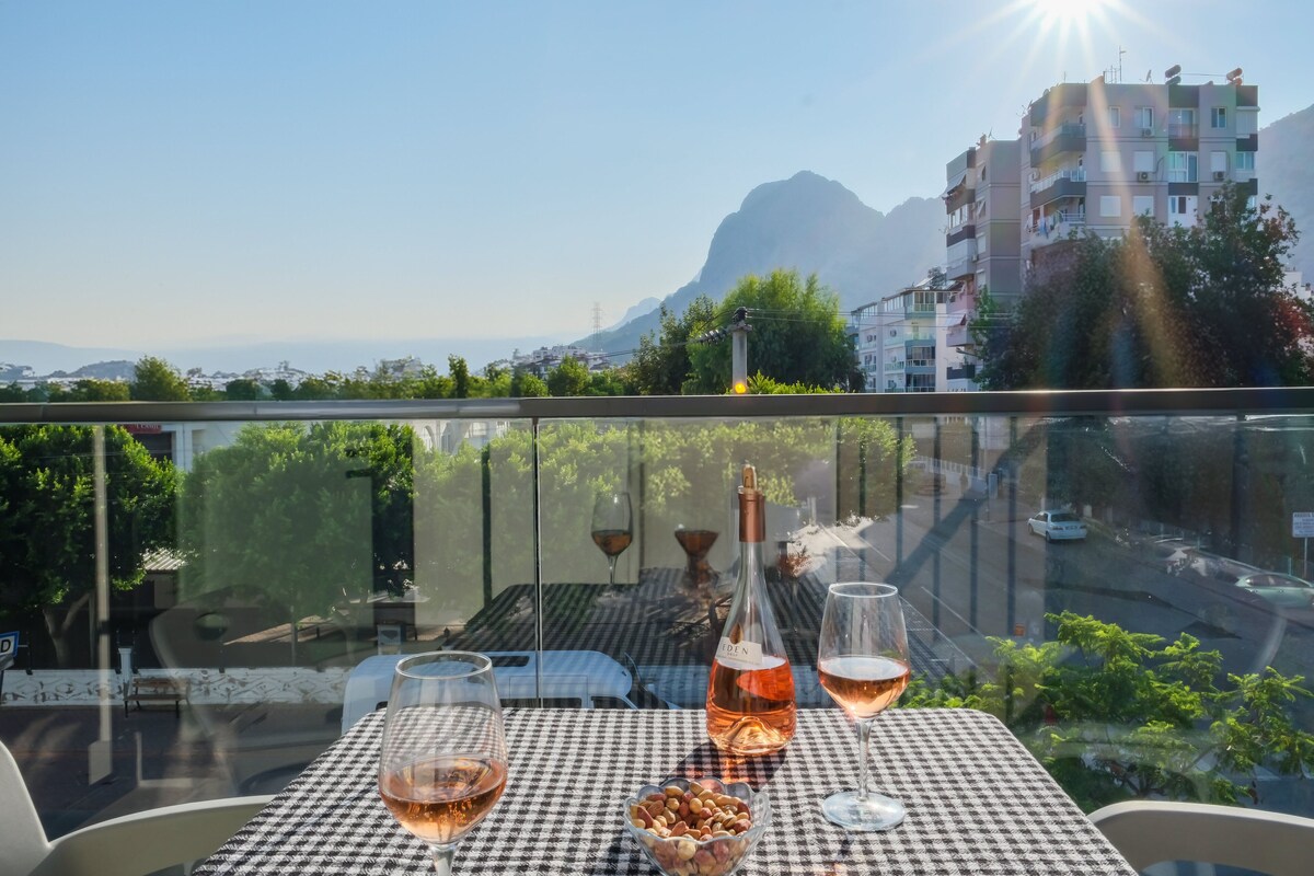 Una mesa con una botella y copas en un balcón con vistas a un paisaje urbano y montañas.