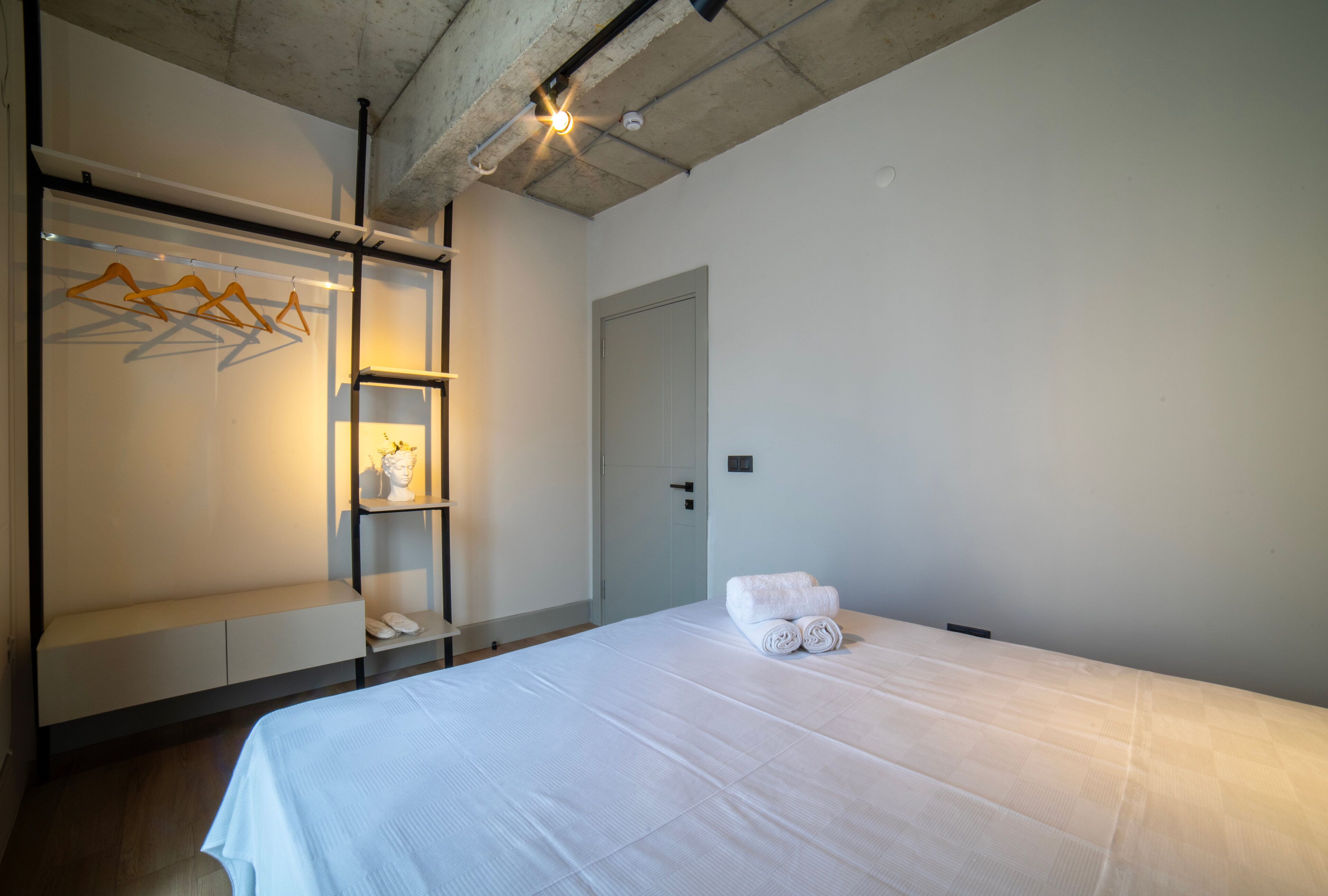 A hotel room with a bed, a wardrobe, a shelf with folded towels, and a door.