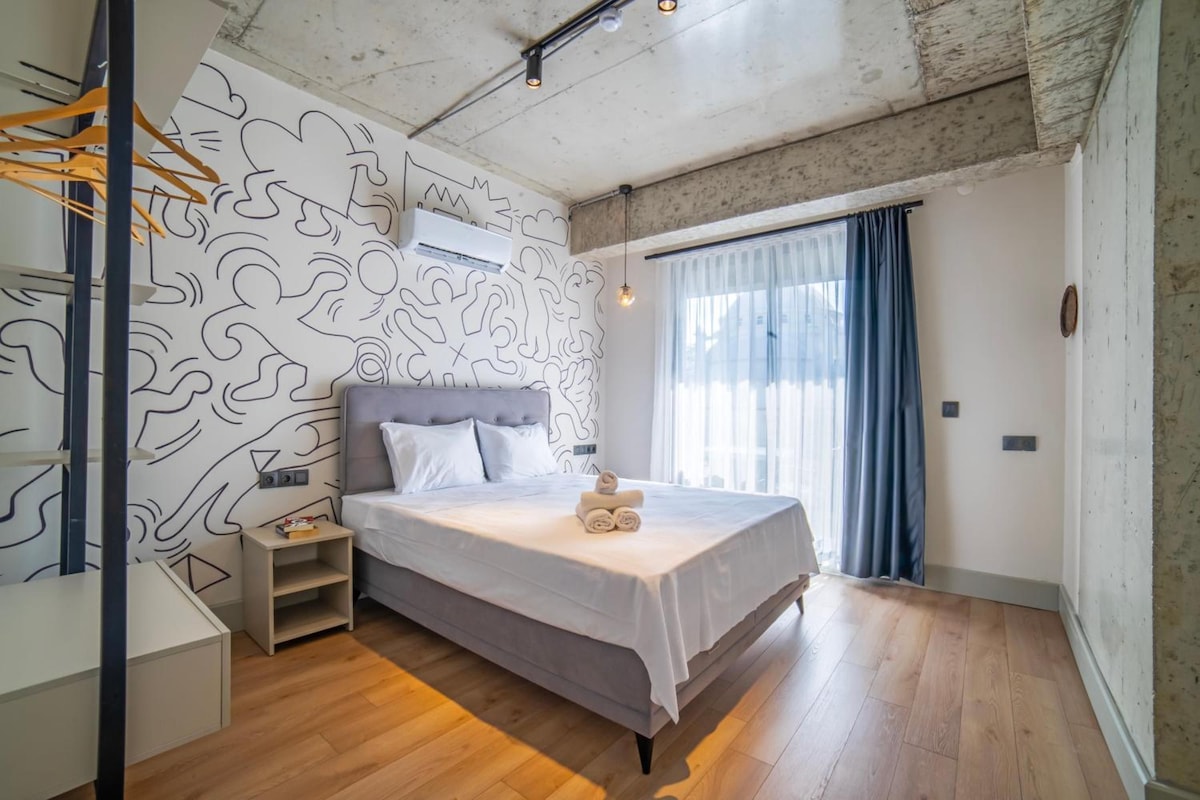 A bedroom with a bed, a nightstand, a wall-mounted air conditioner, and a window with curtains.