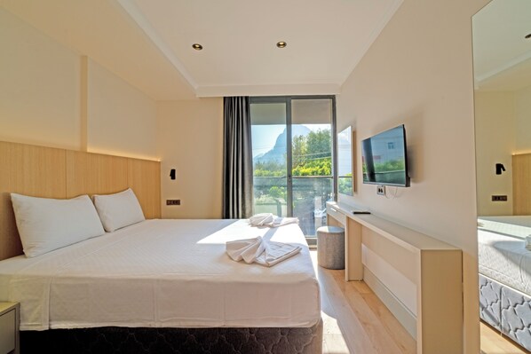 A modern hotel room with a large bed, a television on the wall, and a balcony with a view of greenery and mountains.