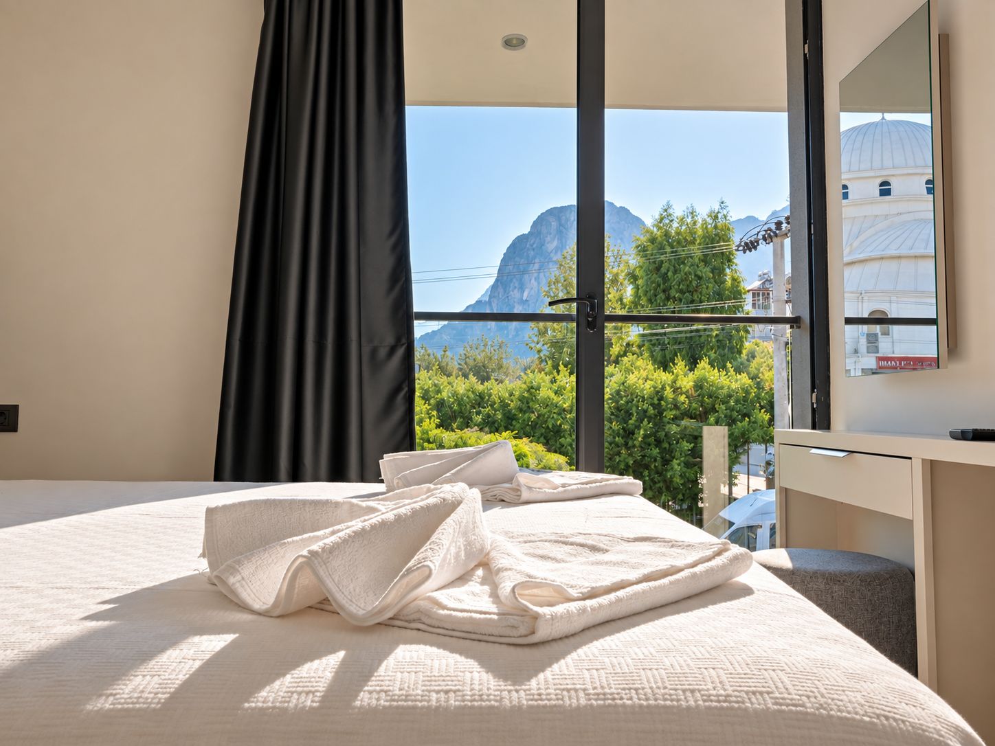 A hotel room with a bed, a window with a view of mountains and greenery, and a desk with a mirror.