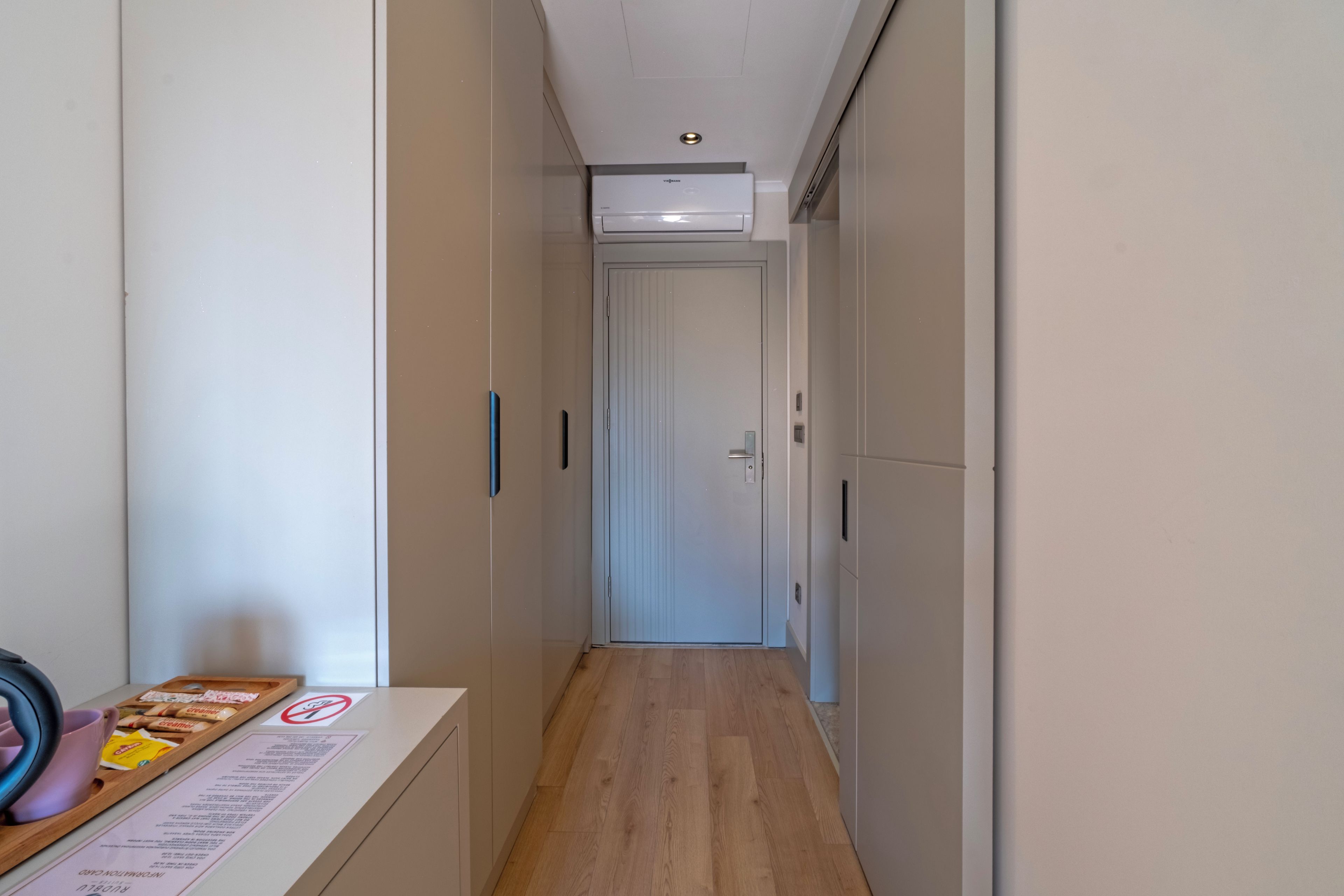 Un pasillo estrecho con suelo de madera, una puerta blanca al final y una pequeña mesa con una bandeja de objetos a la izquierda.