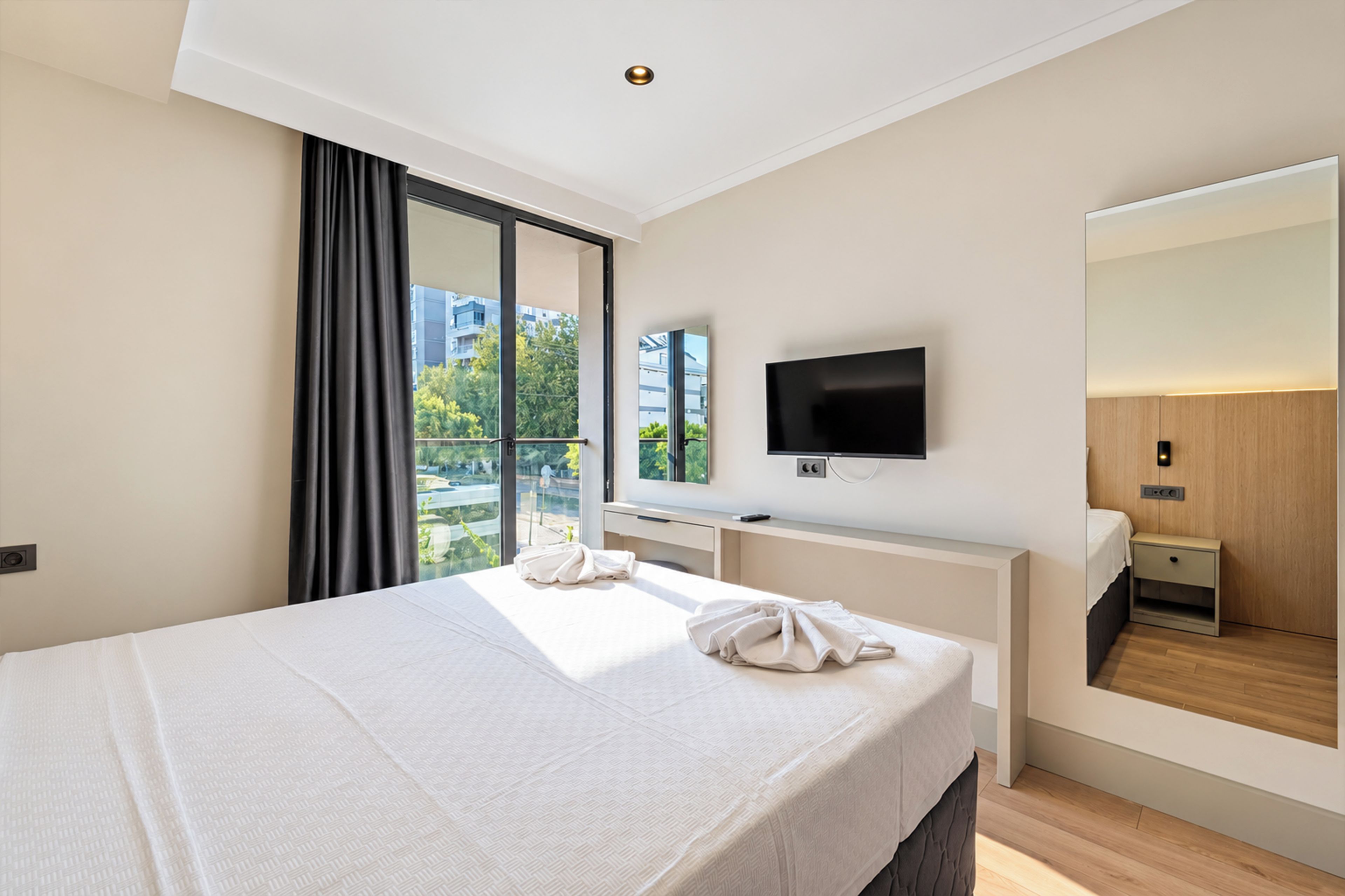 A modern hotel room with a large bed, a flat-screen TV, a large mirror, and a view of the city through the window.