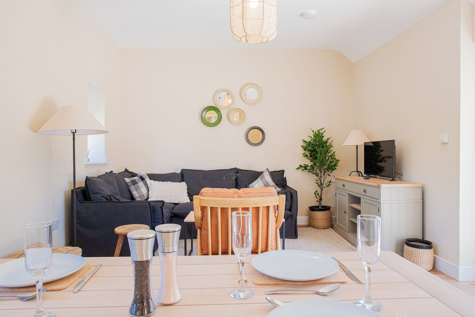A modern living room with a sofa, a dining table set for two, a television, and a small kitchen area.
