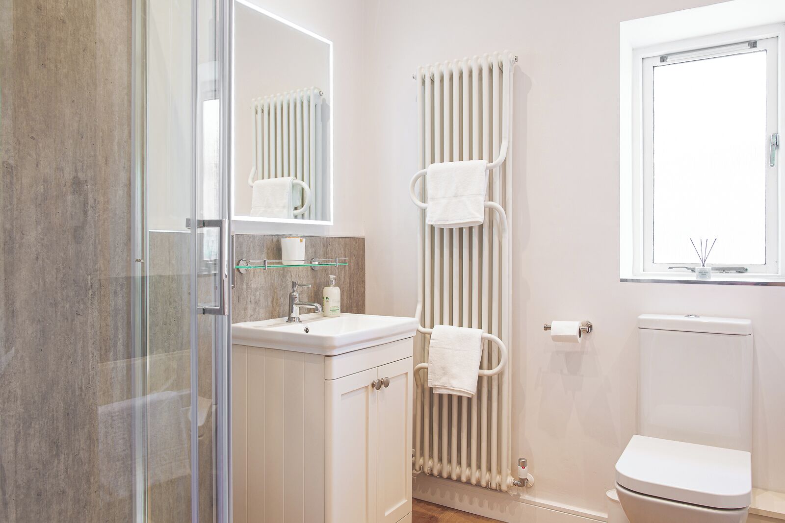 A modern bathroom with a glass shower enclosure, a white sink with a mirror, a towel rack, a radiator, and a toilet.