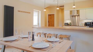 Uma área de refeições com mesa de madeira posta para quatro pessoas, cozinha com eletrodomésticos modernos e uma janela que oferece iluminação natural.