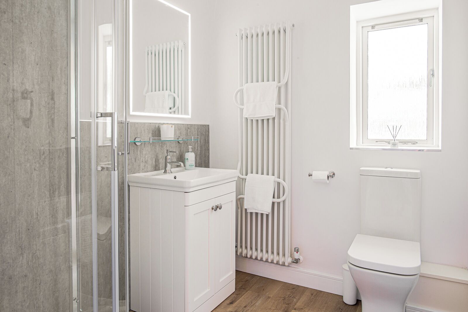 A modern bathroom with a glass shower enclosure, a white sink with a mirror above, a towel rack, a radiator, and a toilet.
