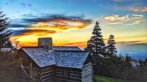 Eine rustikale Holzhütte mit geneigtem Dach, umgeben von dichtem Wald und mit Blick auf ein Gewässer bei Sonnenuntergang.