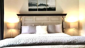 Ein ordentlich bezogenes Bett mit grau-weiß gemustertem Überwurf, einem Holz-Kopfteil und zwei Nachttischlampen.