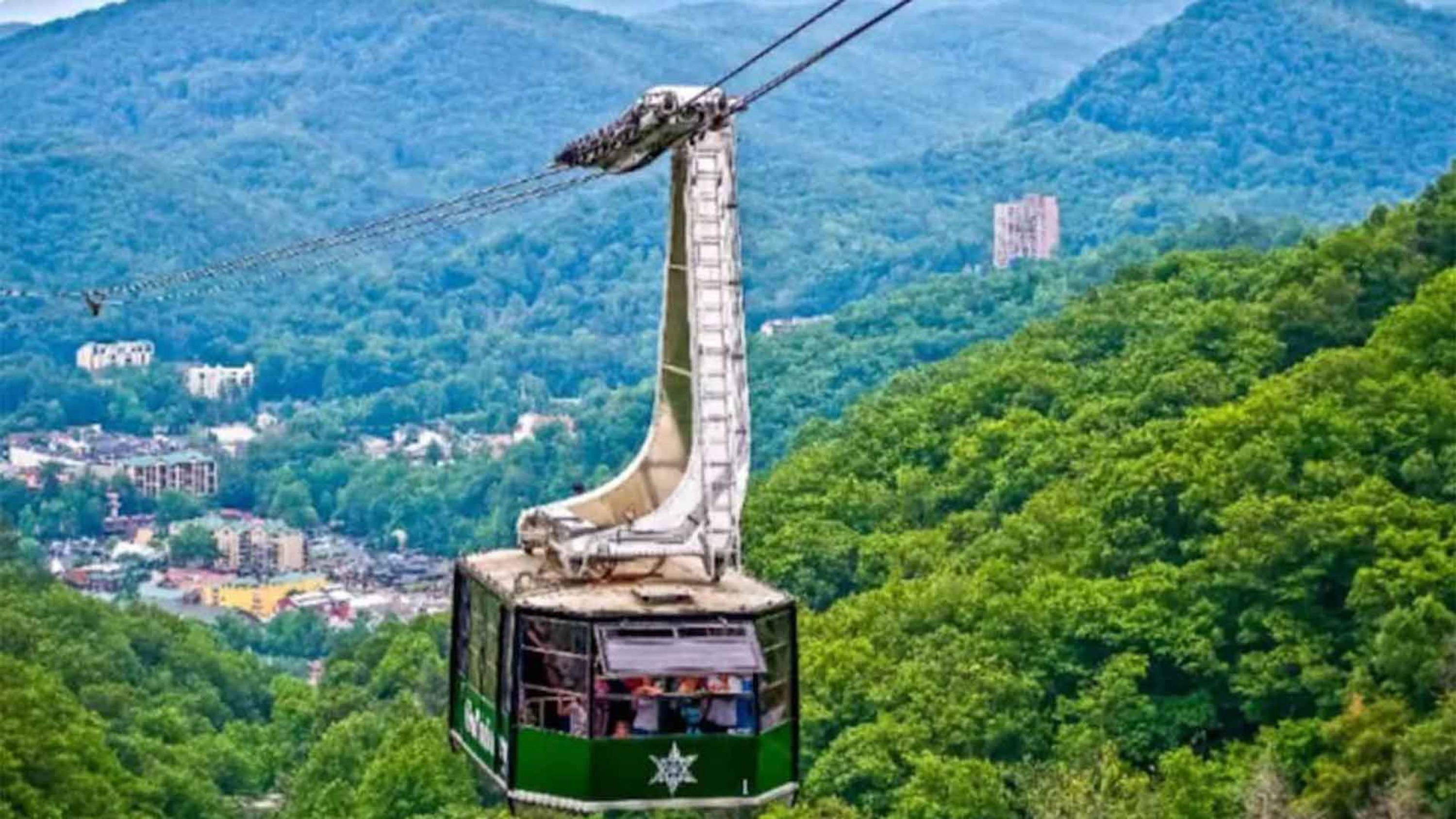A cable car with a green exterior and a star emblem, suspended by cables against a backdrop of lush green hills and scattered buildings.