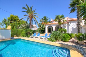 Relax by the serene pool surrounded by lush greenery and palm trees, with a tiled deck and blue lounge chairs.