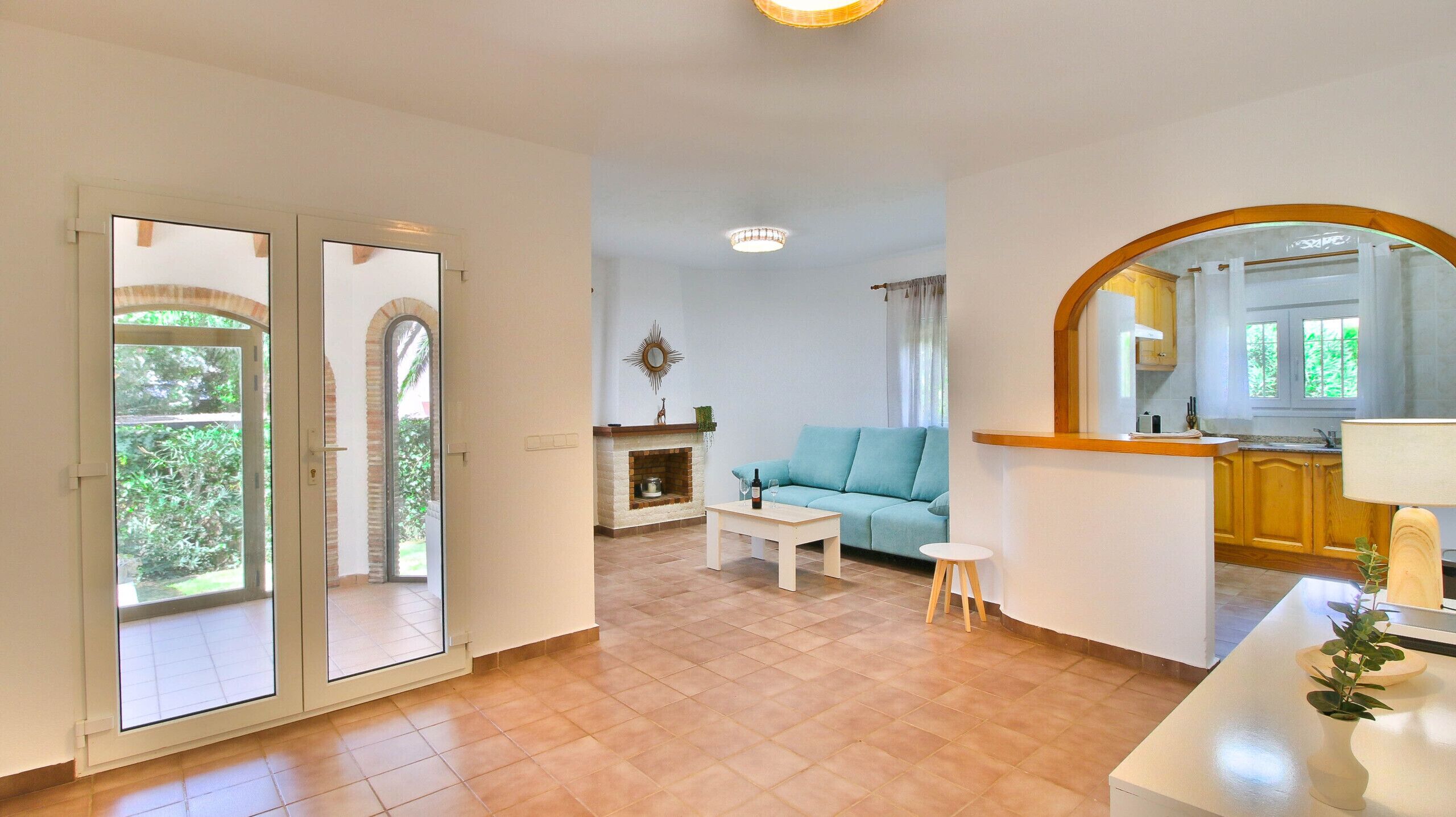 A spacious living area with a sofa, a small table, and a fireplace. There is a kitchen area with a large mirror and a window.
