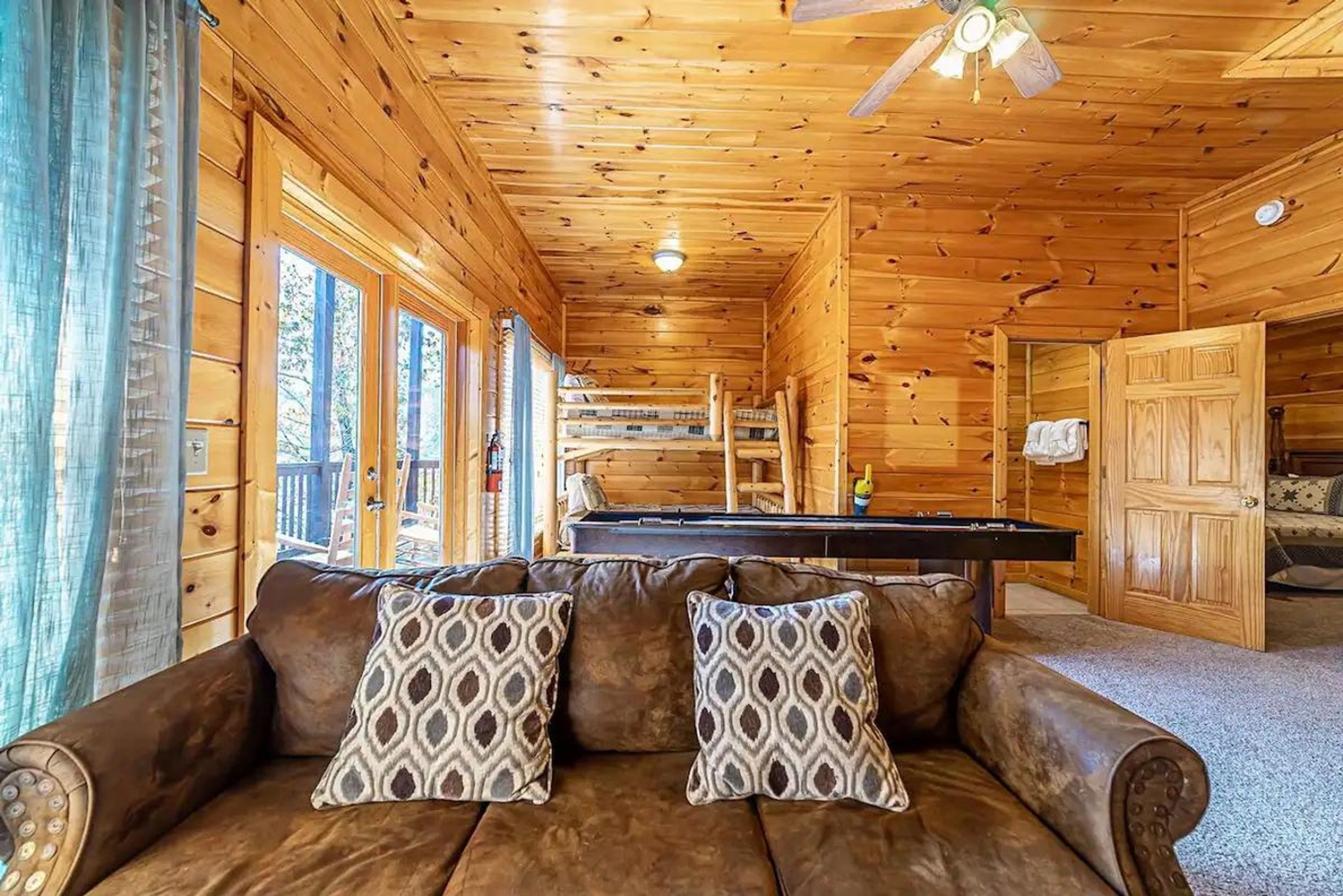 L’intérieur d’un chalet en bois, avec un canapé, des coussins à motifs, une table de billard et un ventilateur de plafond.