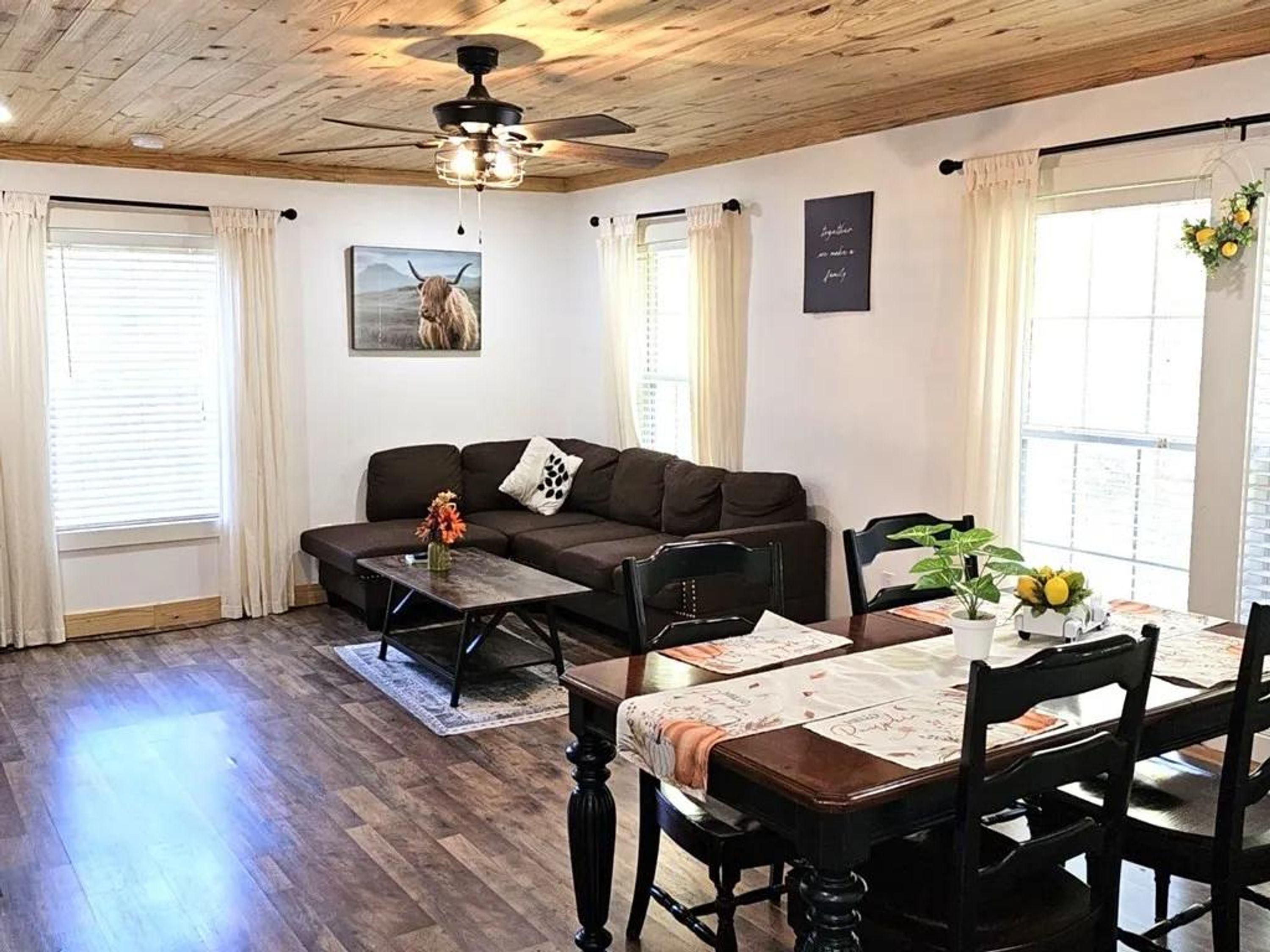 Un salon chaleureux avec un canapé marron foncé, une table basse en bois et une table à manger avec des chaises. On y trouve un ventilateur de plafond, un tableau représentant une vache et une fenêtre avec des rideaux.