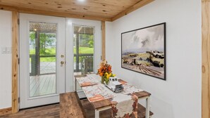 Une pièce avec un plafond en bois, une porte blanche donnant sur l’extérieur, une table recouverte d’une nappe à motifs floraux, un vase rempli de fleurs et un tableau encadré accroché au mur.